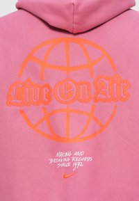 Rozā sporta krekls ar spilgtām oranžām grafikām: "Live On Air" ar globusa dizainu un zemāk baltā krāsā teksts "Making and Breaking Records Since 1972."