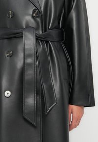 Svart skinn trench coat med dobbeltspent design, store knapper, knyttet belte og glatt, blank overflate.