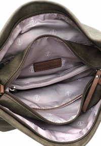 Olivengrüne Canvas-Tasche mit einem hellrosa Innenfutter, ausgestattet mit einem Reißverschlussfach und einem markierten Lederschild. Mehrere Fächer sind sichtbar.
