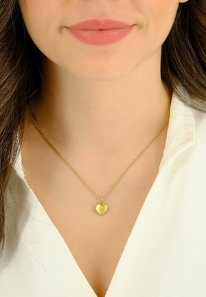 Collana con pendente a forma di cuore in oro su una catena sottile, con una superficie texture e una piccola pietra d'accento. Indossata sopra una camicia bianca con colletto.