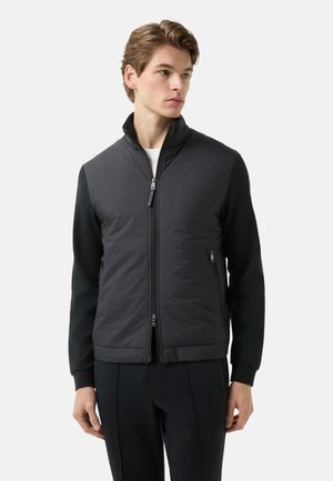 Man draagt een zwart jack met ritssluiting, gewatteerde voorkant en gebreide mouwen, staat voor een effen lichte achtergrond en kijkt opzij.