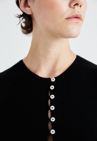 Zwarte gebreide kledingstuk met een ronde halslijn en vijf witte knopen, voorzien van een knoopsluiting, met een gladde textuur.