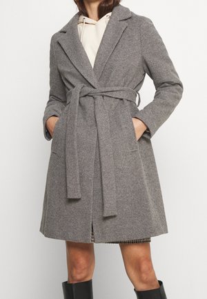 Femme portant un manteau en laine gris ceinturé jusqu'aux genoux, par-dessus un sweat à capuche clair, les mains dans les poches et des bottes noires montant jusqu'aux genoux.