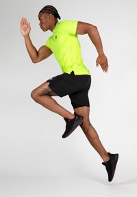 Neon dzeltena sporta krekls ar īsām piedurknēm, saskaņots ar melniem šortiem. Melni skriešanas apavi ar sarkaniem akcentiem, demonstrējot dinamisku pozu.