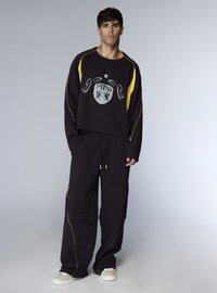 Felpa nera con dettagli gialli e logo, abbinata a pantaloni neri coordinati con cuciture gialle e una vestibilità comoda. Scarpe bianche incluse.