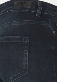 Jeans de ganga azul escuro com uma visão aproximada do bolso traseiro e da cintura. Apresenta um rótulo preto e detalhes de costura sutis.