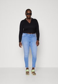 Black long-sleeve top with a collar, light blue slim-fit jeans, and green heeled sandals. Notable silver chain necklace and sunglasses.