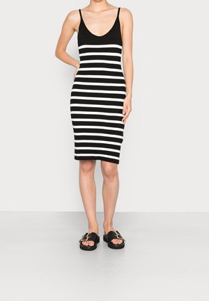 Frau trägt ein ärmelloses Midikleid mit schwarzen und weißen horizontalen Streifen sowie schwarze Slide-Sandalen mit goldenen Schnallen.