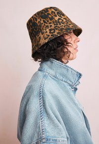 Un chapeau seau avec un motif imprimé léopard dans des nuances de marron et de noir. Le chapeau a une texture de tissu douce et un large bord arrondi.