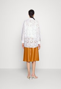 Blouse blanche brodée avec des motifs floraux découpés, associée à une jupe en cuir beige. Texture lisse, coupe ample, manches longues et finition brillante.