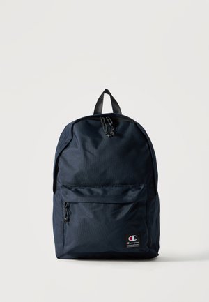 Mochila azul oscuro con un bolsillo frontal con cremallera, asa superior y parche visible con el logo de la marca Champion en la esquina inferior derecha.