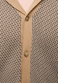 Cardigan tricoté beige et marron à motifs avec deux boutons attachés sur la patte de boutonnage devant.
