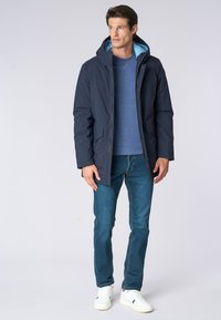 Giacca puffer blu navy con cappuccio, presenta un rivestimento azzurro, due tasche frontali e una chiusura a bottoni, abbinata a jeans blu e sneakers bianche.