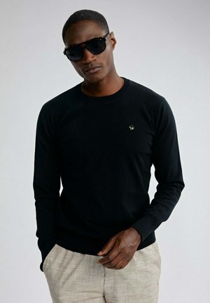 Homme portant une chemise noire à manches longues avec un petit logo, un pantalon beige et des lunettes de soleil noires, sur un fond clair uni.