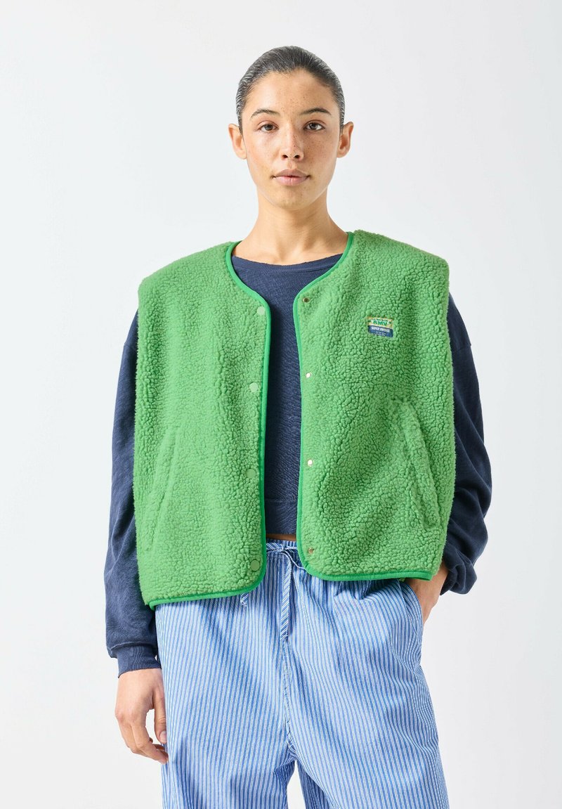 Junge Person mit zurückgebundenem Haar, die eine grüne Fleeceweste über einem dunkelblauen Langarmshirt und blau gestreiften Hosen trägt, vor weißem Hintergrund stehend.