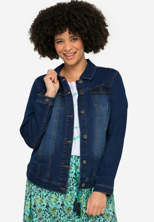 Chaqueta de mezclilla azul oscuro con mangas largas, cierre de botones, dos bolsillos en el pecho y un ligero desvanecimiento. Combinada con una falda floral.
