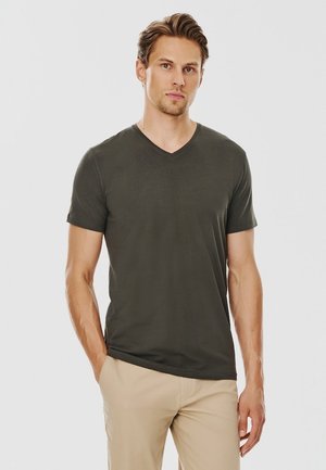 Ciemnozielony t-shirt z dekoltem w serek, krótki rękaw, gładka tekstura, lekki materiał, w połączeniu z beżowymi spodniami, model stoi na białym tle.