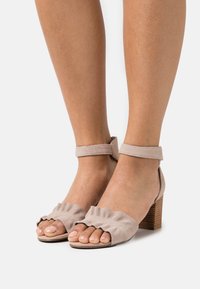 Beżowe zamszowe sandały na obcasie z paskiem wokół kostki, zdobione falbaną na palcach, z drewnianym słupkowym obcasem, prezentujące gładką teksturę i otwarty fason na palce.