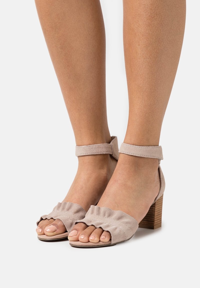 Beżowe zamszowe sandały na obcasie z paskiem wokół kostki, zdobione falbaną na palcach, z drewnianym słupkowym obcasem, prezentujące gładką teksturę i otwarty fason na palce.