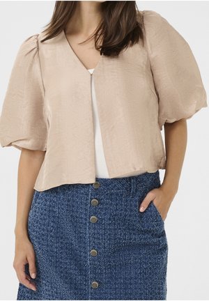 Vrouw draagt een beige cropped blouse met ballonmouwen en een blauwe, hooggetailleerde, getextureerde rok met knopen aan de voorkant en zakken.