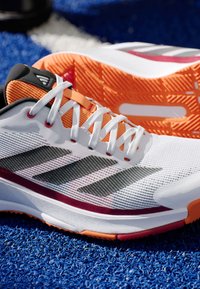Witte sport schoenen met grijze strepen, oranje accenten en een gestructureerde zool. Beschikt over een lichtgewicht mesh bovenwerk en een traditioneel vetersysteem.