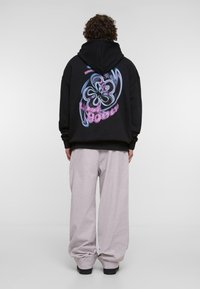 Melnā kapucēta svītra ar krāsainu grafiku aizmugurē, apvienota ar gaiši pelēkiem platām biksēm un melniem apaviem.