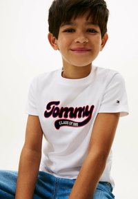 T-shirt bianca a maniche corte con bold navy e rosso con la scritta "Tommy" e "CLASS OF 1995" sottostante, abbinata a jeans in denim blu.