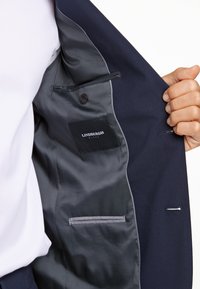 Wnętrze marynarki w kolorze granatowym ma gładką satynową podszewkę, wewnętrzną kieszeń z guzikiem oraz tłoczoną metkę. Elegancki dopasowany design z szarymi akcentami.