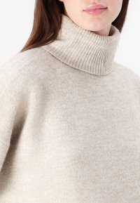 Beige høyhalset genser laget av mykt stoff, med ribbestrikket krage og en løs passform med subtil tekstur.