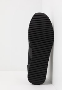 Suela de zapatilla negra con surcos horizontales apoyada contra una esquina blanca de la pared y el suelo.