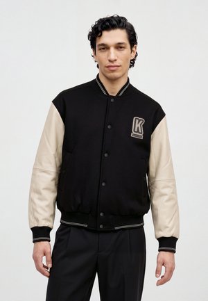 Un homme porte une veste universitaire noire à manches crème, avec des bordures rayées et un écusson portant la lettre K, sur un fond clair et uni.