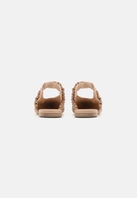 Sandales slip-on beiges avec un design de sangle froncée, semelle plate et dos ouvert. Le matériau semble doux et lisse. Style simple et minimaliste.