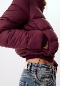 Burgundy pufferjakke med quiltet tekstur, ribbede manchetter og en justerbar kant, båret over lyseblå denimjeans.