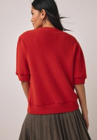 Rdeč teksturiran pulover s kratkimi rokavi z rebrastimi manšetami in robom. Model ga nosi čez naborkano karo krilo. Mehka, rahlo puhasta tkanina.