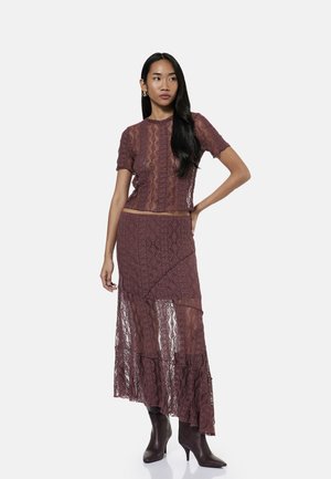 Femme aux longs cheveux noirs porte un top en dentelle violette transparente et une jupe asymétrique assortie avec des bottes marron à talons pointus, debout avec la main sur la hanche.