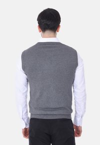 Maglione grigio lavorato a maglia senza maniche, con orlo a coste, indossato sopra una camicia bianca a maniche lunghe, con dettagli di cucitura visibili sulle spalle e sul collo.