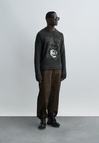 Hombre con gafas de sol oscuras, suéter negro con diseño de serpiente y calavera, pantalones marrones sueltos y botas oscuras con cordones, de pie frente a un fondo liso.