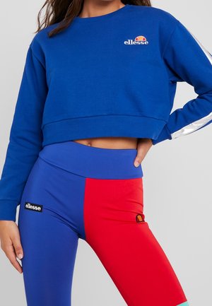 Vrouw draagt een blauwe, korte ellesse sweater en kleurige leggings in blauw en rood met ellesse-logo's op beide kledingstukken.