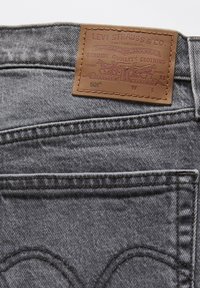 Gray denim jeans with a brown leather label featuring embossed text and imagery. The design includes curved stitching on the back pockets.