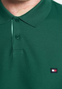 Gros plan sur une personne portant un polo vert avec deux boutons attachés et un petit logo rectangulaire rouge, blanc et bleu sur la poitrine.