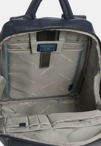 Interno di uno zaino in pelle blu con fodera grigia, scomparti imbottiti e dettagli di marca. Visibile tasca con zip e spallacci regolabili.