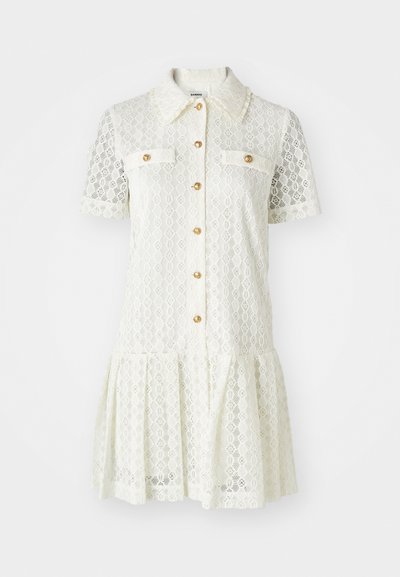 Robe en dentelle crème avec des manches courtes, un col, des boutons dorés et deux poches poitrine. Jupe évasée avec un détail en bordure scallopée.