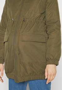Parka de couleur vert olive avec des poignets élastiques, une taille cintrée et deux poches plaquées à l'avant. Fabriquée en tissu lisse et léger.