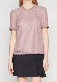 Femme portant un top court à manches courtes en dentelle florale mauve avec des bords festonnés et une mini jupe plissée noire.