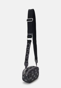 Bolso cruzado de tela negra con cierre de cremallera, que presenta un patrón de logotipos repetidos y una correa ancha con el texto "LULU" en gris.