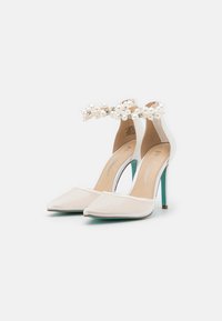 Scarpe bianche con tacchi alti, punta affusolata, sovrapposizione trasparente e una cinghia alla caviglia decorata con perle e strass. Accento verde sul tacco.