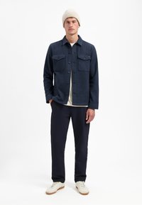 Marineblauwe button-up jas met twee borstzakken, gedragen over een crèmekleurig shirt, gecombineerd met donkere broek en lichte sneakers, aangevuld met een beige beanie.