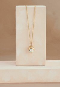 Kuldne kaelakee, millel on pärlpendant, mis on kujundatud näo sarnaselt ja sellel on kaks väikest silma ning kuldse aktsendiga juuksekujundus. Tekstuuriga neutraalne taust.