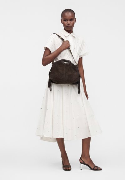 Femme en robe blanche tenant un grand sac à main en daim marron foncé avec des pampilles, portant des sandales à talons hauts noires à bout ouvert.