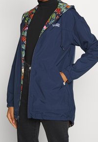 Granatowa lekka kurtka z kwiatową podszewką, regulowanym kapturem ze ściągaczem, bocznymi kieszeniami oraz logo Vans na piersi.